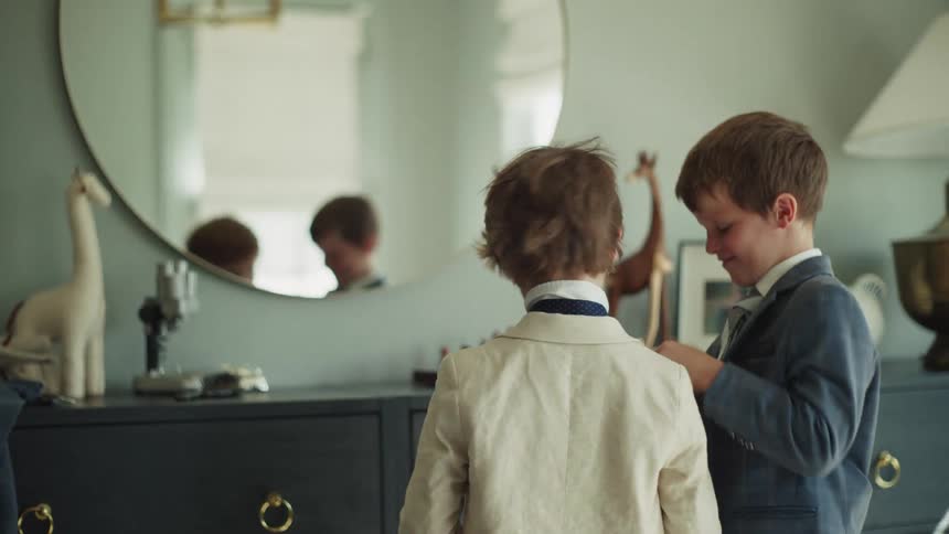 A young boy smiles as he helps his younger brother adjust his suit jacket while getting ready in front of a mirror
