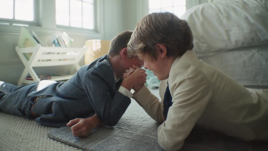 Two young boys dressed in suits playfully arm wrestle on the floor of a sunlit bedroom