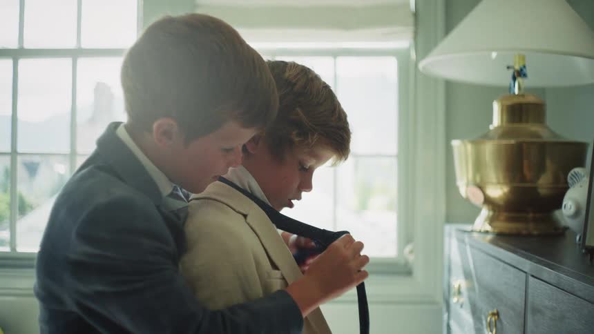 An older brother carefully helps his younger brother put on his necktie in a sunlit bedroom