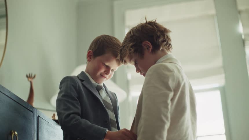 A young boy lovingly helps his brother get dressed in a formal suit and tie in their bedroom