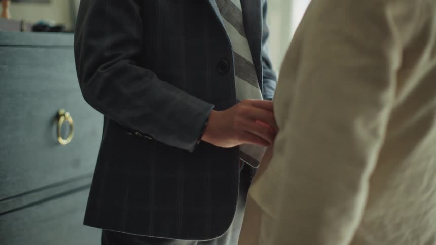 A close-up of a young boy in a suit and tie carefully adjusting his brother's jacket in their room