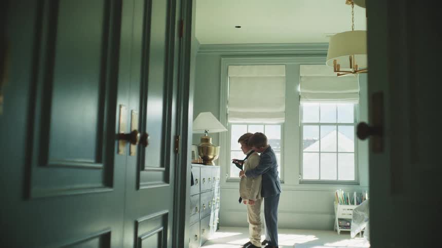 An older brother helps his younger sibling with his suit and tie in a beautifully lit bedroom before a formal event
