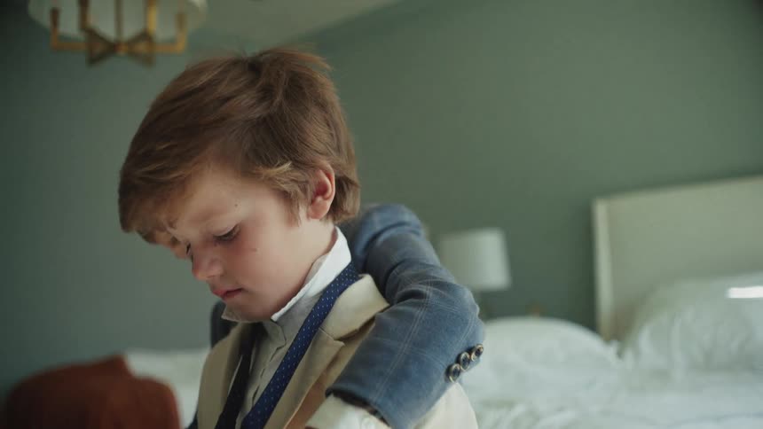 An older brother helps his younger sibling get ready for a formal event, carefully adjusting his tie in a softly lit bedroom