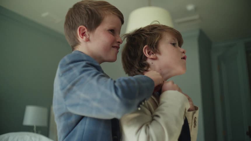 A heartwarming moment as one young brother carefully helps the other with his suit and tie while getting ready in a bedroom
