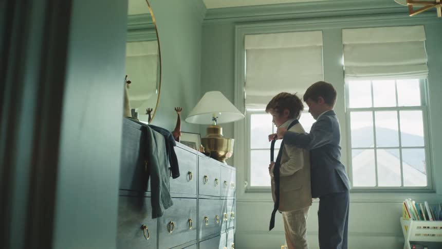 A young boy helps his little brother put on a tie as they get ready in their sunlit bedroom