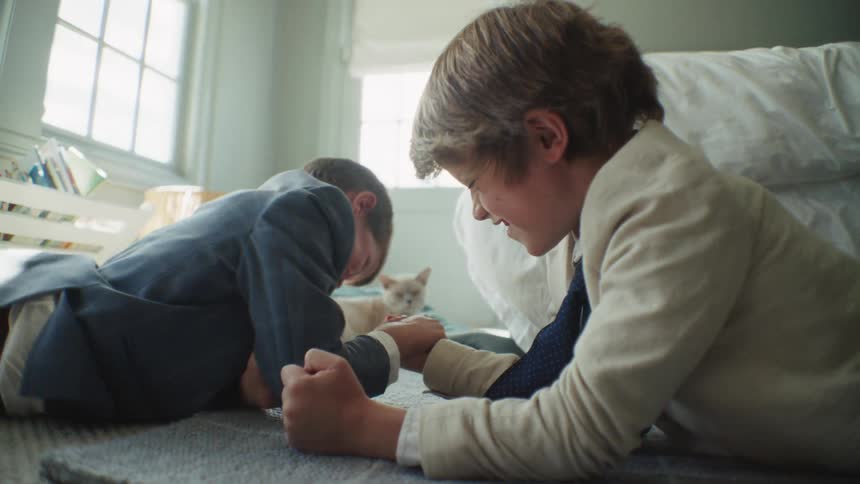 Two young brothers in suits laugh and smile while playfully arm wrestling in slow motion on their bedroom floor