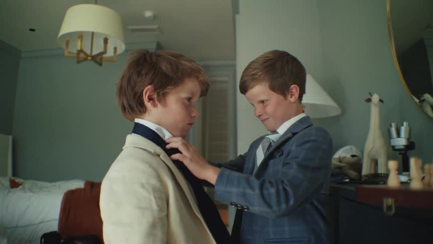 In a heartwarming slow-motion shot, an older brother carefully adjusts his younger brother's tie as they prepare for a formal event