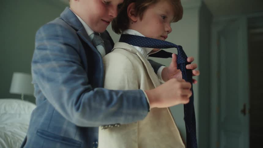 A caring older brother helps his younger brother tie his necktie as they get dressed in formal suits in slow motion