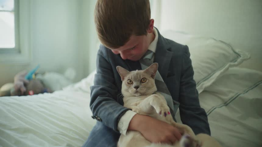 A dapper young boy in a suit lovingly holds his cat with painted nails while sitting on his bed