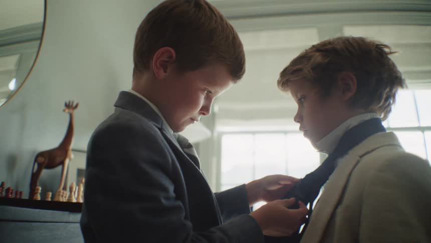 A caring older brother helps his younger brother tie a necktie as they get ready in their suits