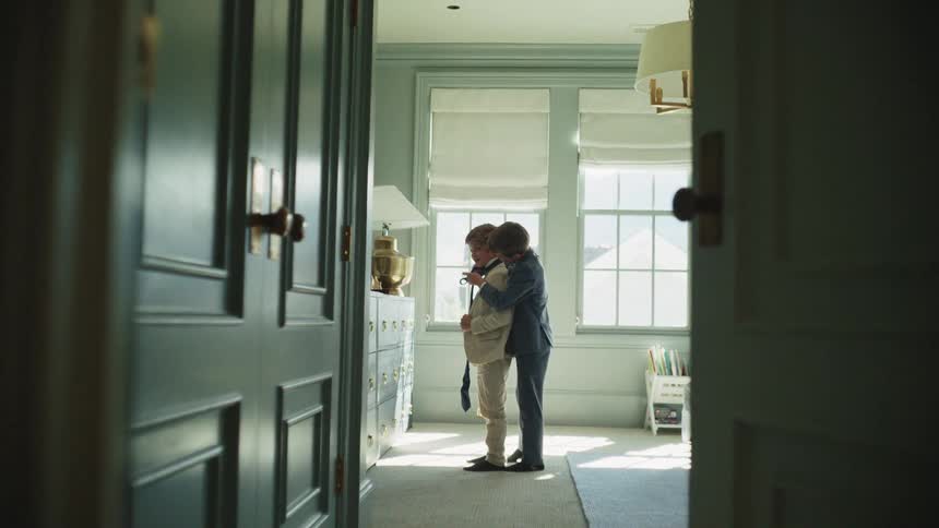 A young boy helps his brother put on a tie while they get dressed in formal suits in a sunlit bedroom