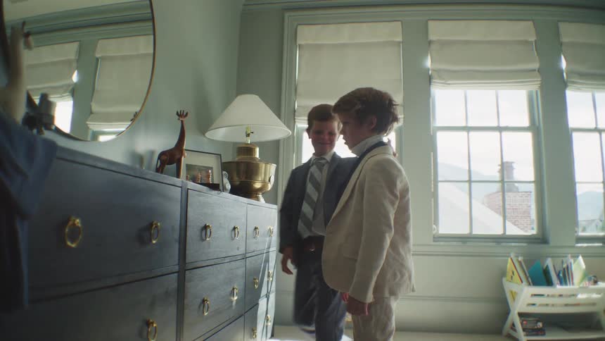 An older brother helps his younger brother tie his tie as they get ready together in their sunlit bedroom