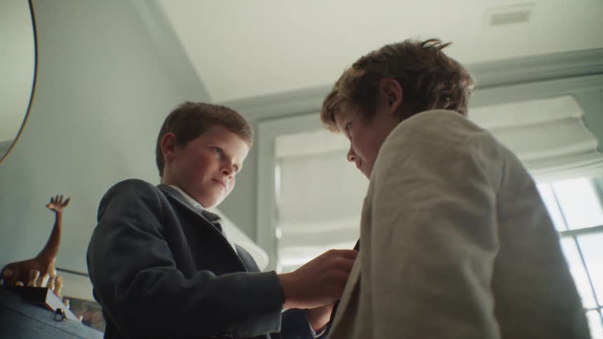 A young boy smiles proudly as he helps his brother get ready, carefully tying his necktie for him in their bedroom
