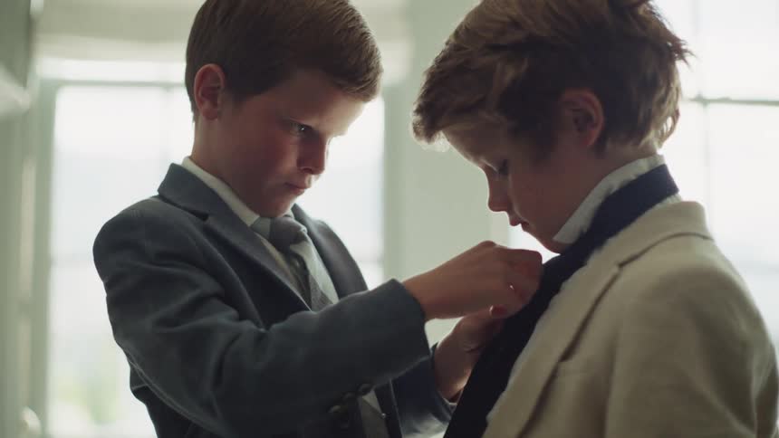 A young boy carefully helps his brother adjust his necktie as they get ready for a formal event in a sunlit room
