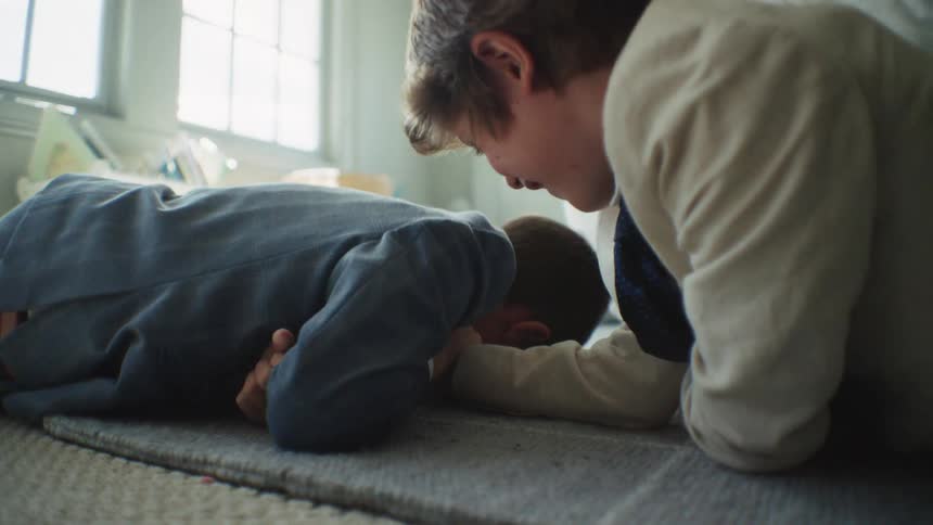 Two smiling brothers dressed in suits have a playful arm-wrestling match on the floor in slow motion, with one eventually winning