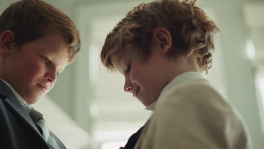 A close-up of a young boy kindly helping his friend adjust his necktie as they get dressed in formal suits