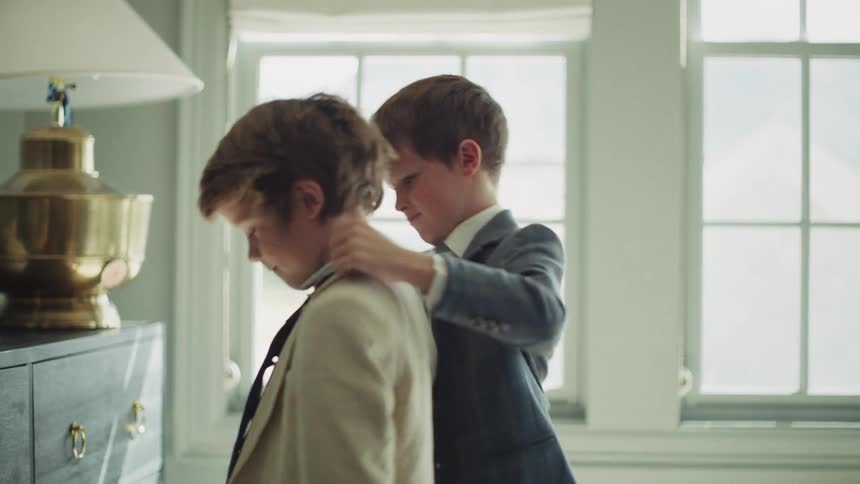 A young boy helps his older brother adjust his suit and tie as they get ready together in a sunlit room