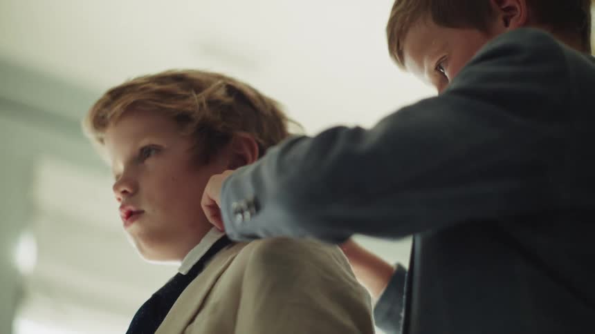 A caring young boy helps his brother adjust his suit and tie as they get ready together indoors