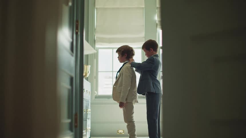 A young boy helps his brother adjust his suit and tie as they get ready for a formal event in a beautiful, sunlit room