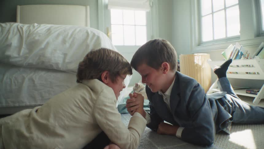 Two laughing young boys in suits playfully arm wrestle on the floor of a sunlit bedroom