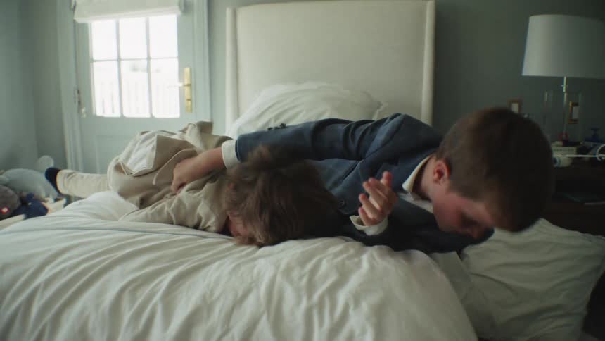Two young brothers wearing suits playfully wrestle and jump on a bed in slow motion, laughing with joy in their sunlit bedroom