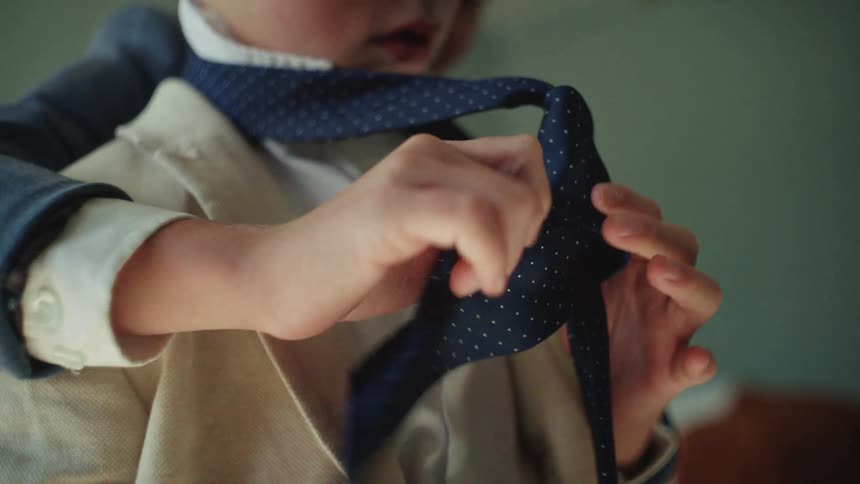A close-up slow motion shot of a young boy patiently helping his brother tie his necktie as they get dressed in suits