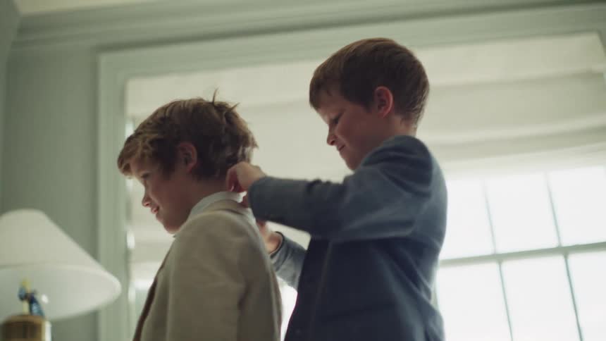 A young boy smiles as he helps his brother adjust his formal suit and collar in a bright, sunlit room