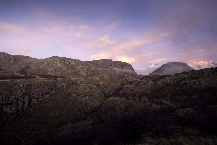 A serene timelapse captures clouds and fog rolling through a rugged mountain valley at dawn, revealing a snow-capped peak in the warm morning light