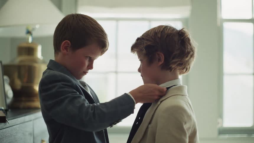A young boy carefully helps his brother adjust his collar and tie as they get ready in their formal suits