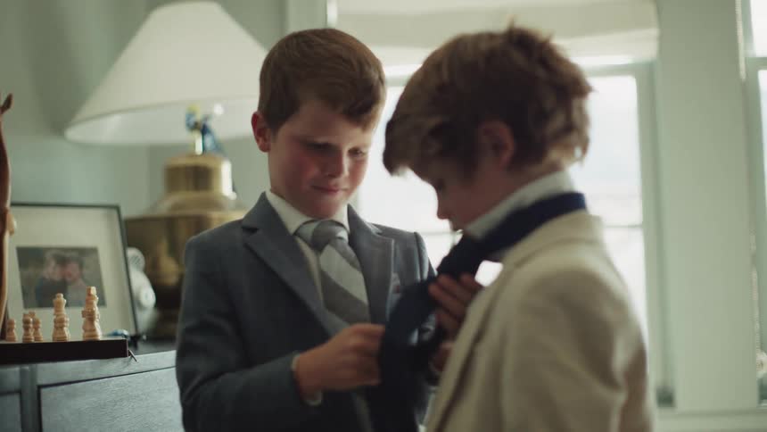 A young boy in a suit carefully helps his brother tie his necktie inside a sunlit room