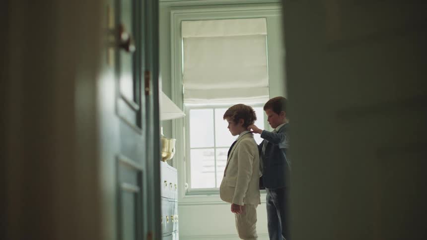 Framed through a doorway, an older brother helps his younger brother get dressed in formal suits for a special occasion