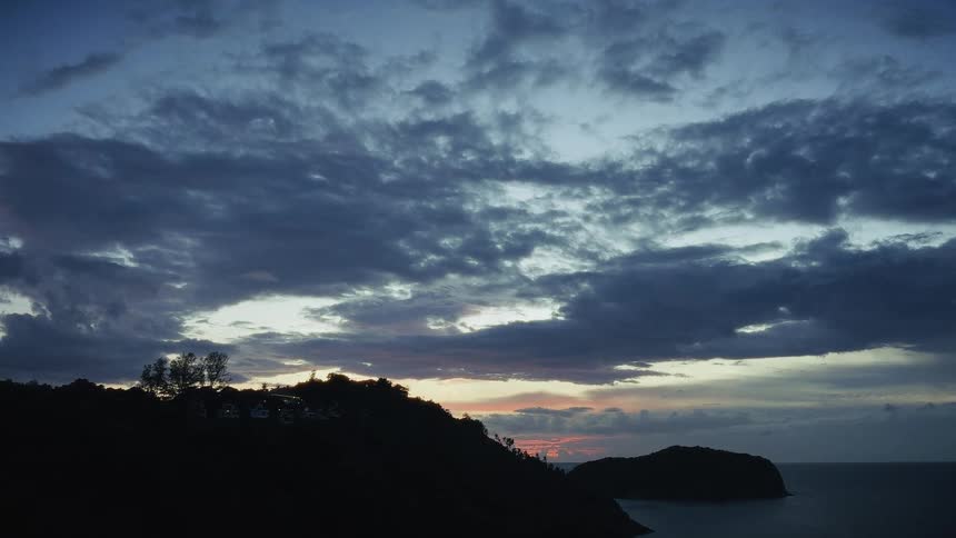 A dramatic cloudy sky looms over a silhouetted coastline with a small island at sunrise