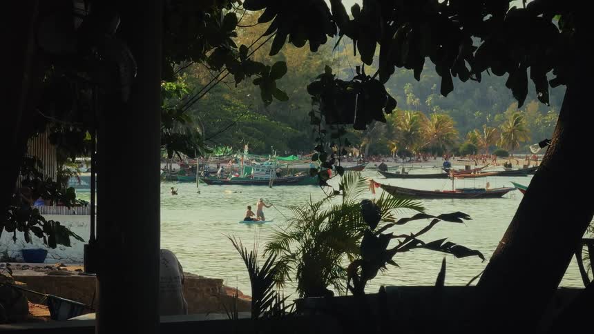 A serene view through silhouetted trees of people paddleboarding in a tropical bay filled with long-tail boats