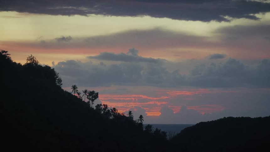 Silhouetted tropical hills with palm trees frame a stunning, colorful sunset over a calm ocean