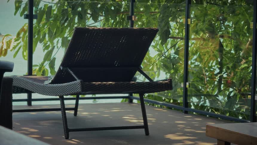 A tranquil static shot of a wicker sun lounger on a modern balcony with a glass railing, overlooking a calm sea and tropical foliage