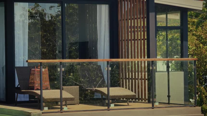 A serene view of a modern hotel balcony with lounge chairs and a birdcage, with lush trees reflected in the glass doors