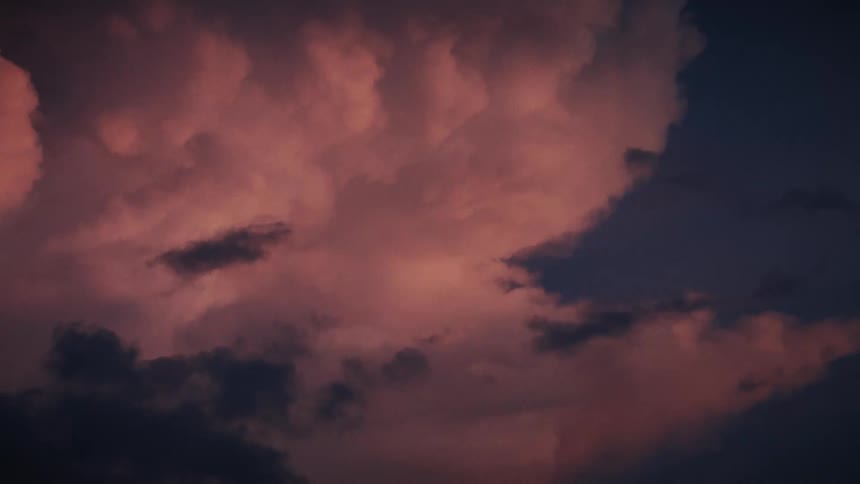 A beautiful sunset casts a pink and purple glow on dramatic storm clouds as they slowly drift and flash with lightning