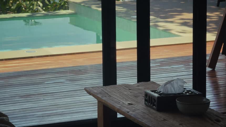 A tranquil view from inside a room shows a rustic table with a tissue box, looking out to a serene swimming pool and wooden deck