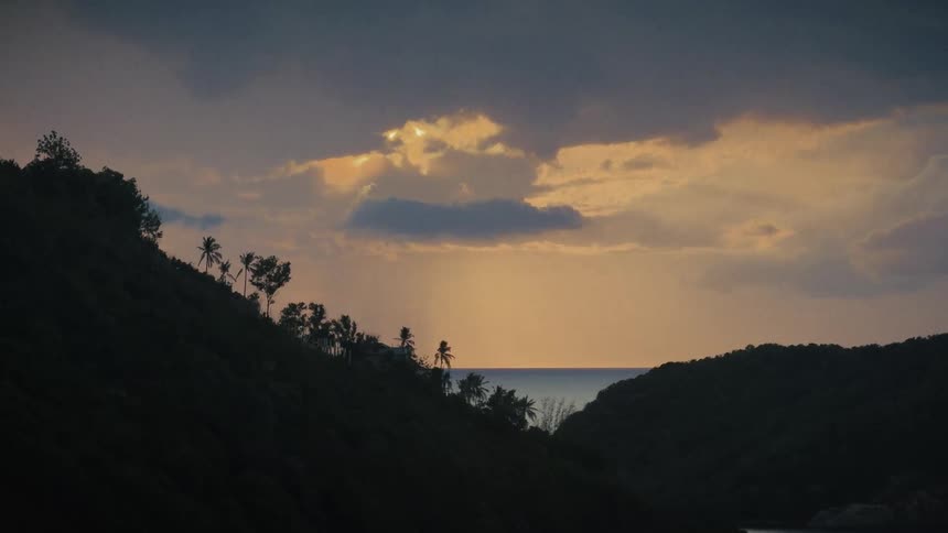 A beautiful tropical sunset over the ocean is seen through a valley between two silhouetted hills