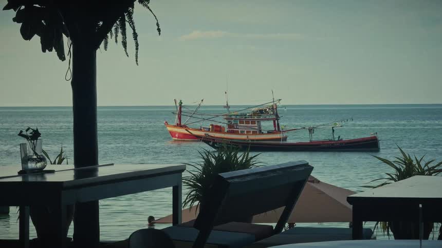 A traditional fishing boat is anchored in the calm ocean as seen from a tropical resort patio