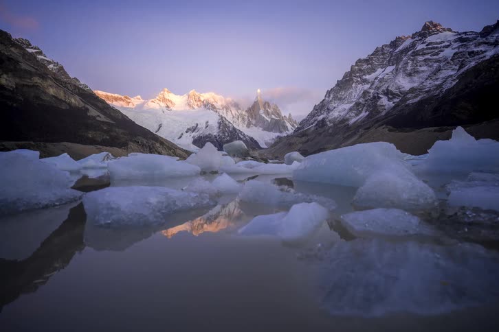 A timelapse captures a serene sunrise over Patagonia's snow-capped Cerro Torre mountain, with icebergs floating in a still glacial lake as the moon sets
