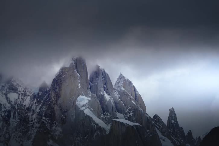 Dark storm clouds move ominously over a jagged, snow-covered mountain range