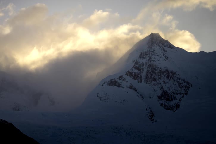 A time-lapse of majestic, snow-covered mountain peaks as golden clouds and fog roll over them