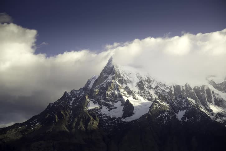 A majestic snow-capped mountain peak is partially obscured by slow-moving clouds under a deep blue sky