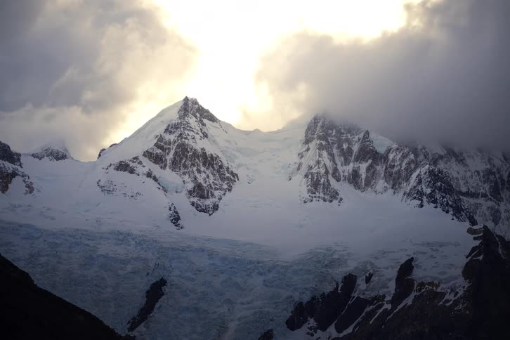 A dramatic, static view of a sunlit, snow-covered mountain peak and glacier shrouded in dark clouds