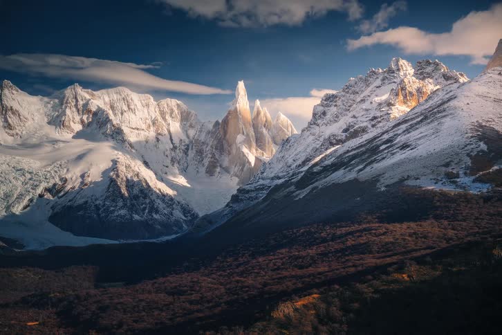 Majestic view of the sunlit, snow-covered Cerro Torre mountain peaks in Patagonia with clouds gently drifting across the deep blue sky