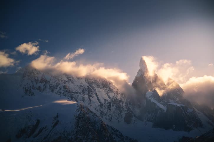 Golden sunlight illuminates dramatic clouds swirling around the majestic snow-capped peaks of a mountain range