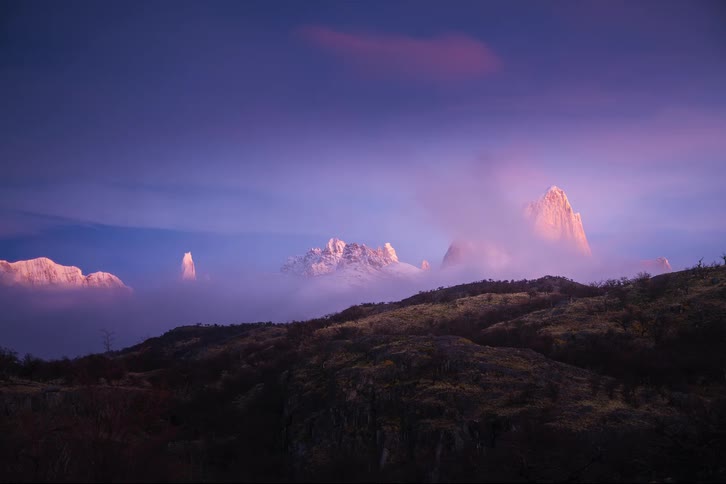 A breathtaking timelapse captures the vibrant purple and pink hues of sunrise illuminating the snow-covered peaks of Mount Fitz Roy