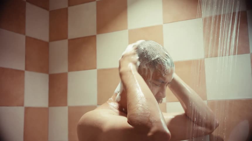 A shirtless man with a shaved head lathers up his head with soap while taking a shower in slow motion