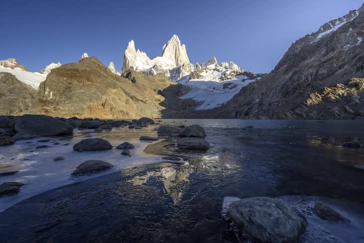 A stunning timelapse of the majestic Mount Fitz Roy reflecting in a partially frozen alpine lake as sunlight moves across the rugged Patagonian landscape